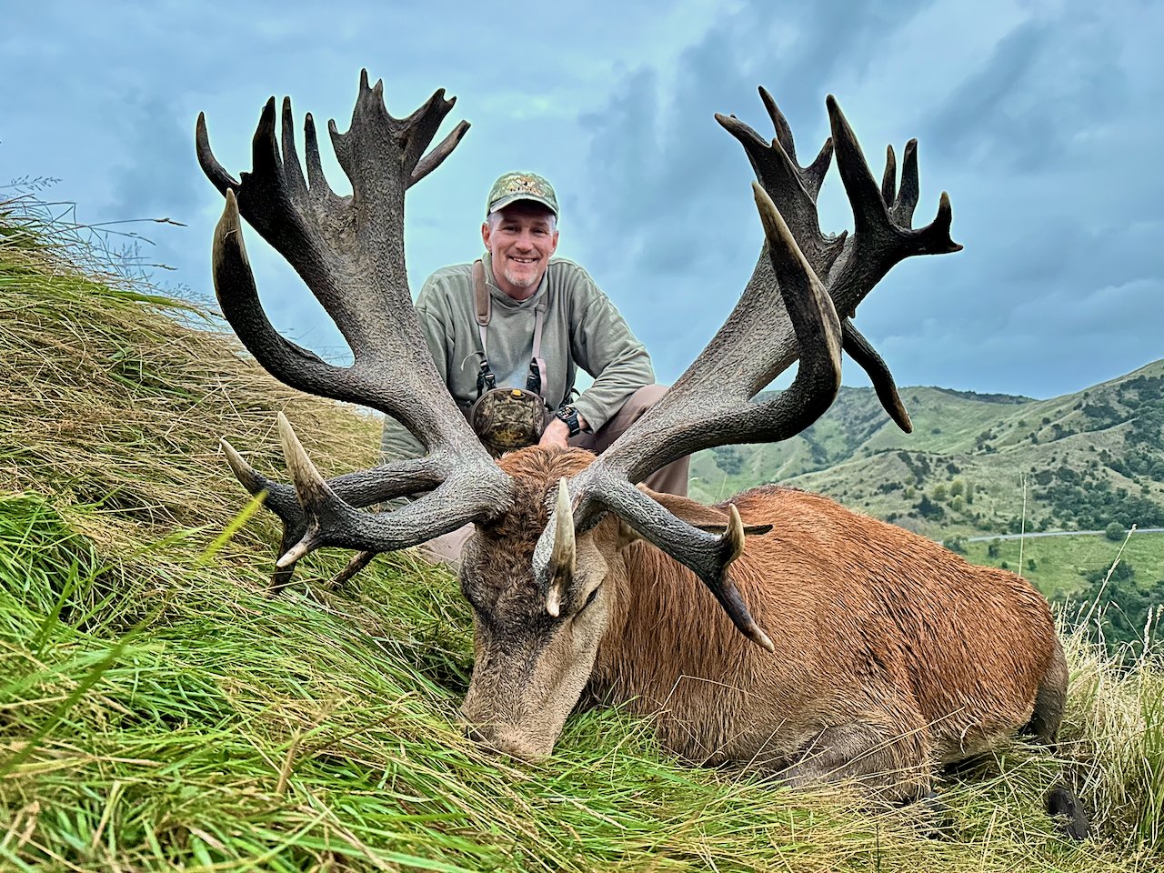 A man in outdoor gear crouches behind a large elk with impressive antlers, lying on green grass in a hilly landscape with cloudy sky.