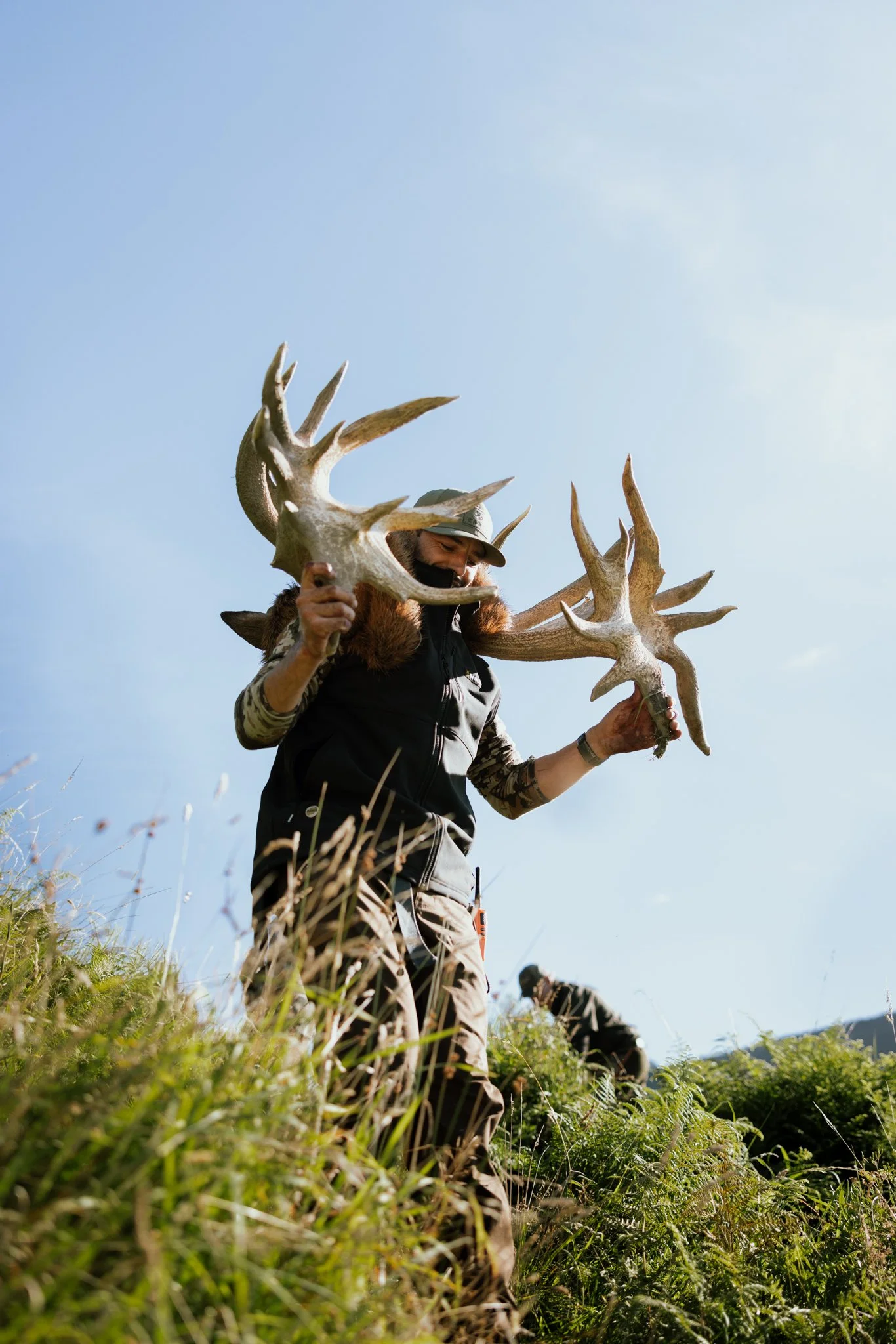 Person holding large antlers outdoors in a grassy field against a blue sky.