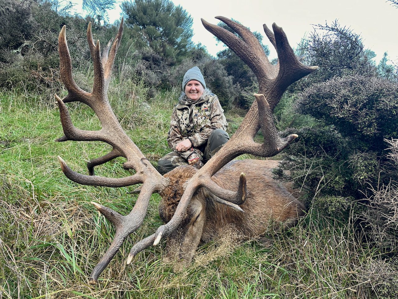 A woman in camouflage clothing and a gray beanie, smiling, kneeling behind a large elk with impressive antlers, in a grassy and bushy outdoor setting.