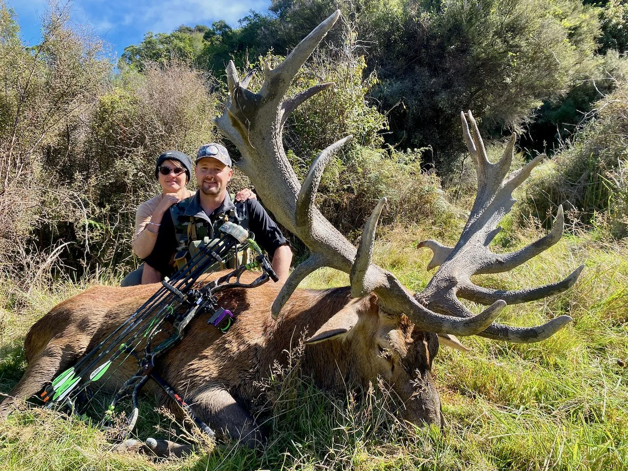 Two people, a woman and a man, smiling and posing next to a large, dead elk with impressive antlers in a grassy, wooded area.