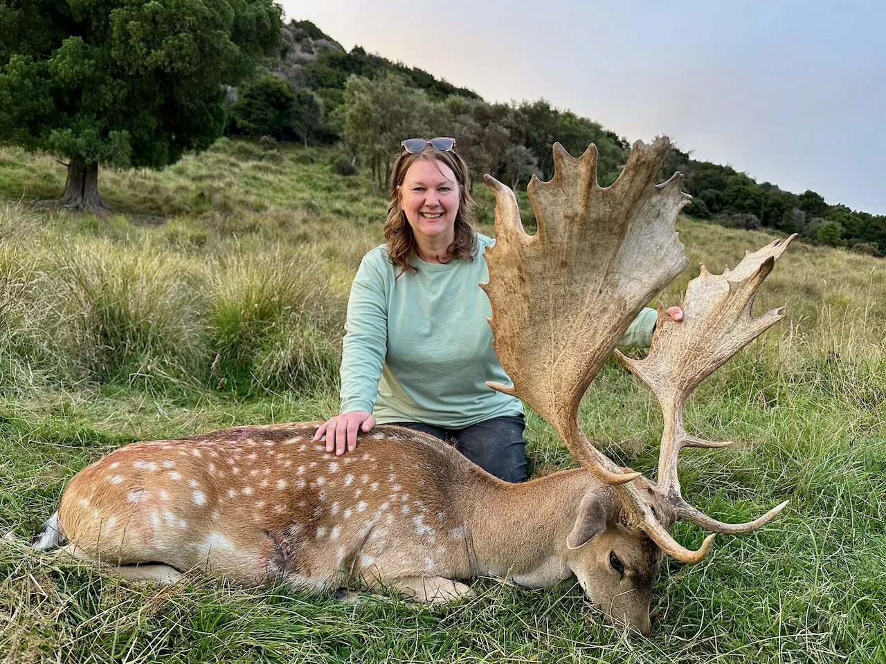 A woman with glasses on her head smiling while sitting next to a dead deer with large antlers in a grassy field.