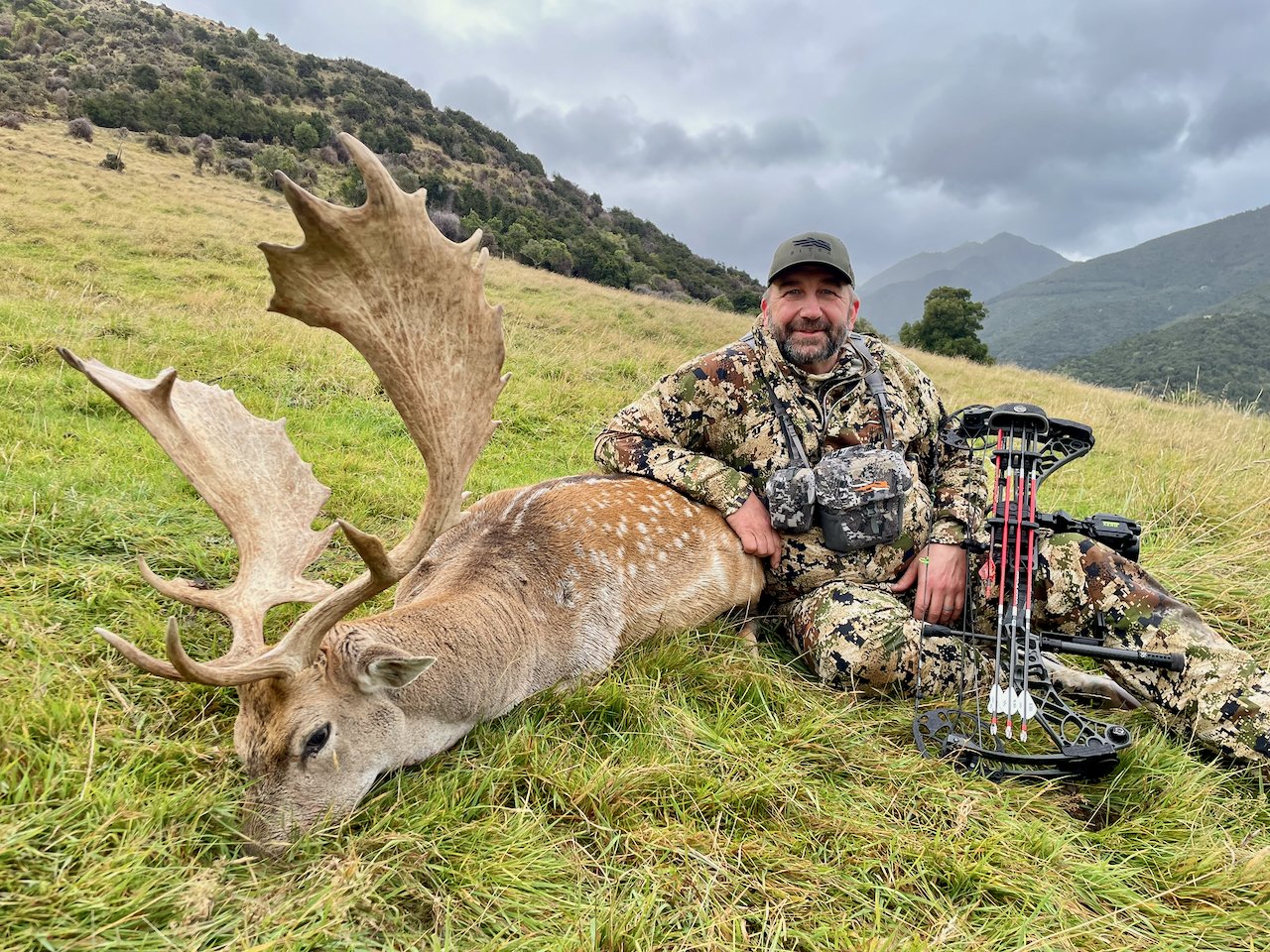 A man in camouflage clothing sitting on the grass next to a large, dead deer with prominent antlers. The man is smiling and has archery equipment lying on the ground nearby. The background features grassy hills and a cloudy sky.
