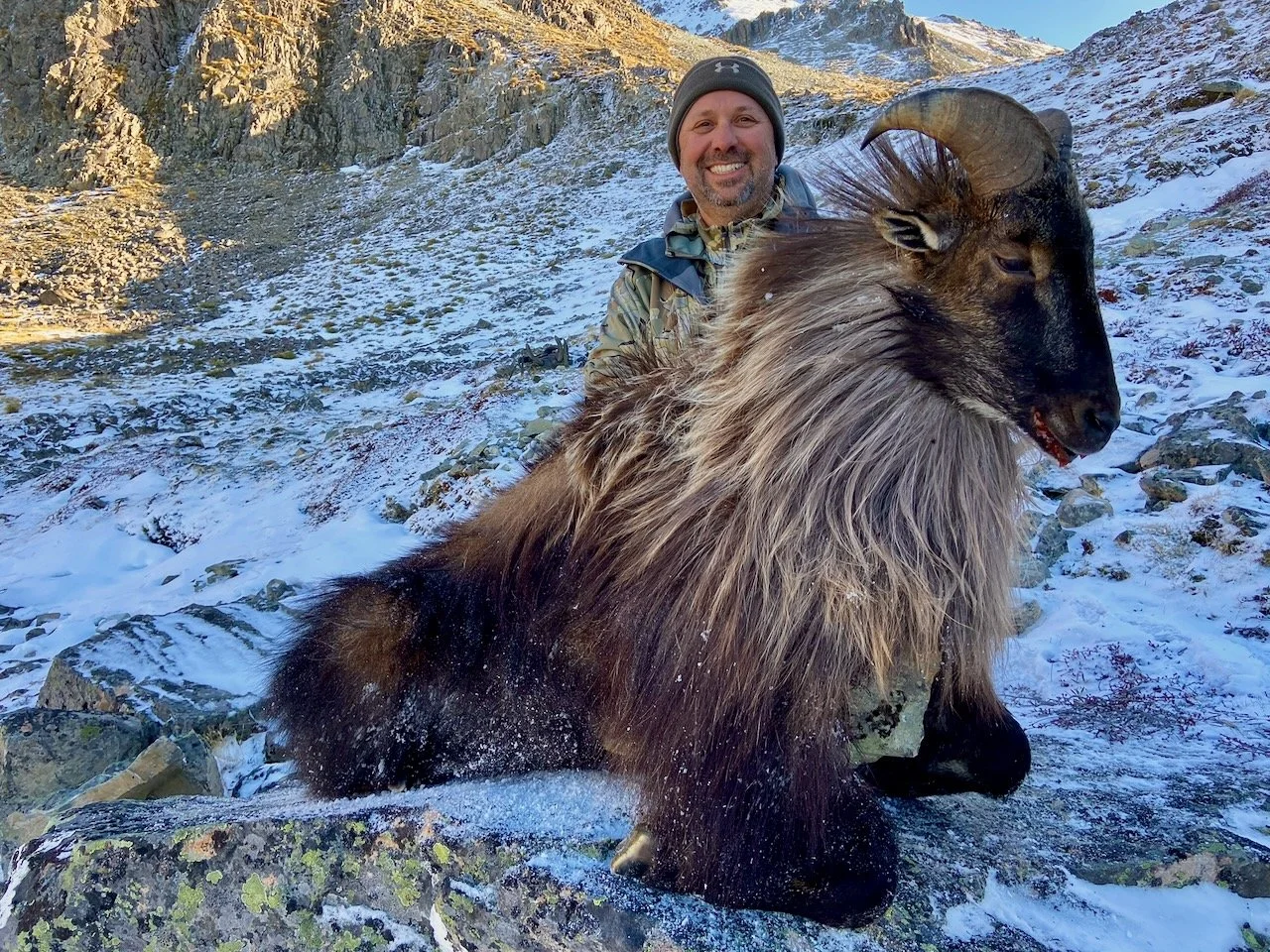 A man in outdoor gear smiling behind a large mountain goat with curved horns, standing on snowy rocky terrain with mountains in the background.