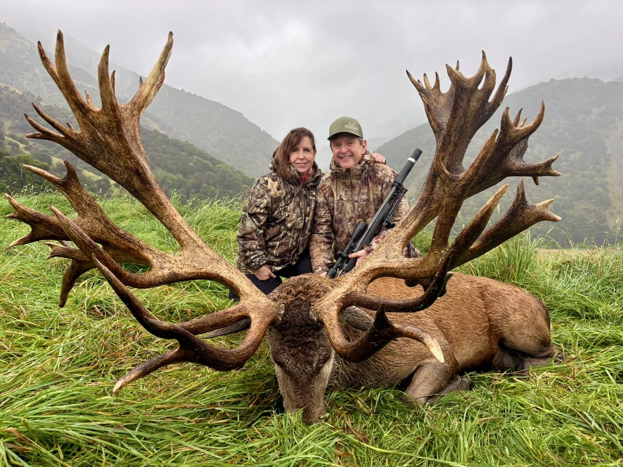 Two hunters in camouflage clothing sitting behind a large deer with big antlers, on a grassy field with mountains in the background. One hunter is holding a rifle.