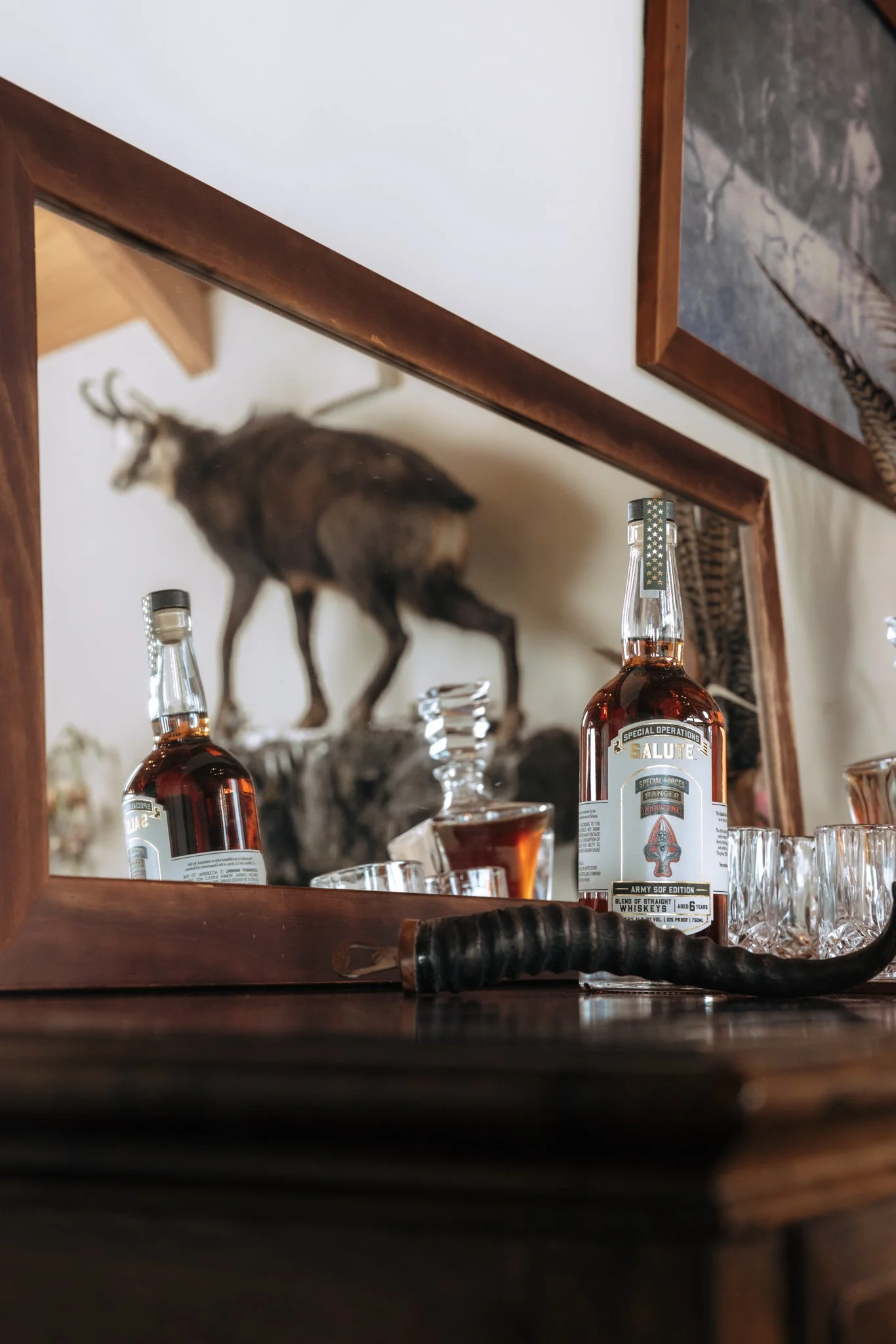 A mirror reflecting a wooden shelf with liquor bottles, a glass of whiskey and a mounted deer head in the background.