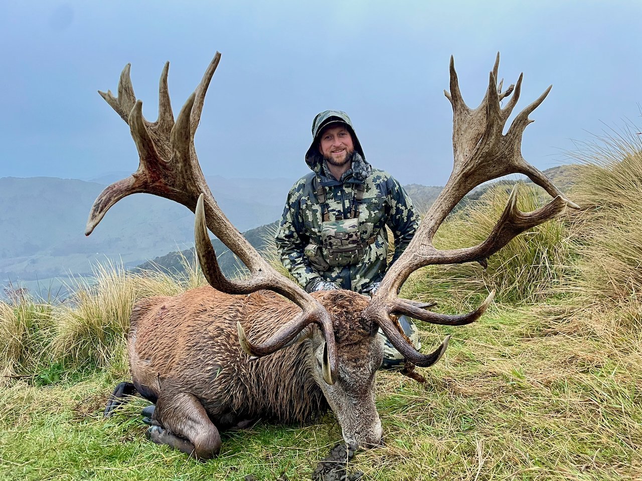 A man in camouflage clothing and hat kneels next to a large hunted stag with massive antlers, on a grassy hillside with mountains in the background.