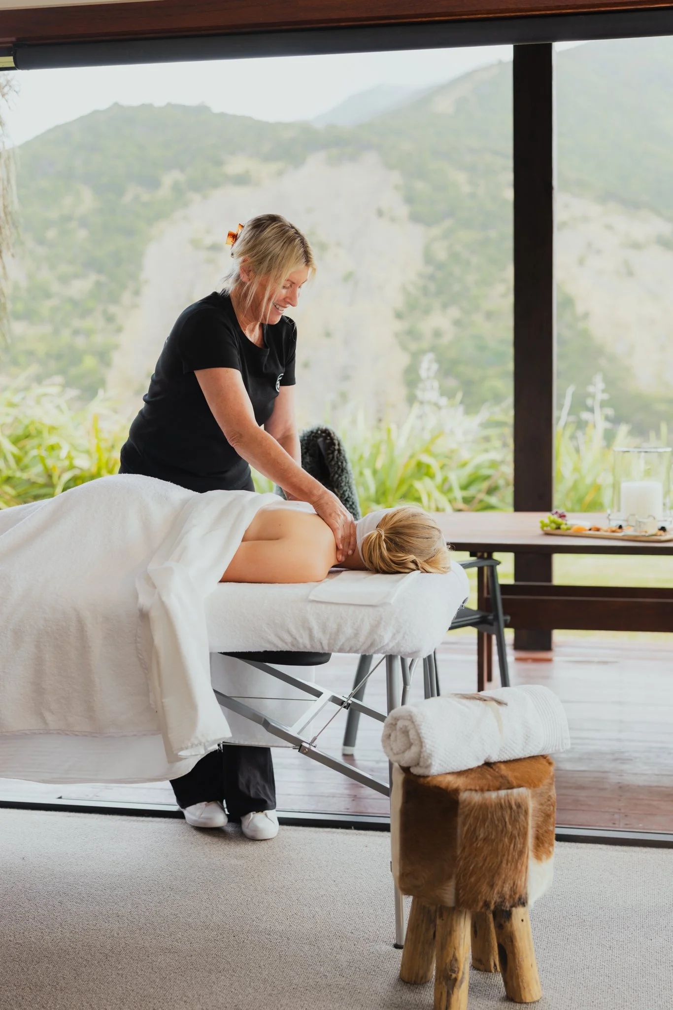 A woman receiving a massage from a masseuse with a natural mountain view outside.