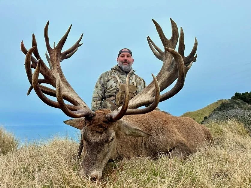 A man in camouflage clothing and a backward cap stands behind a large elk with prominent antlers lying on grass in a natural landscape with hills and a cloudy sky
