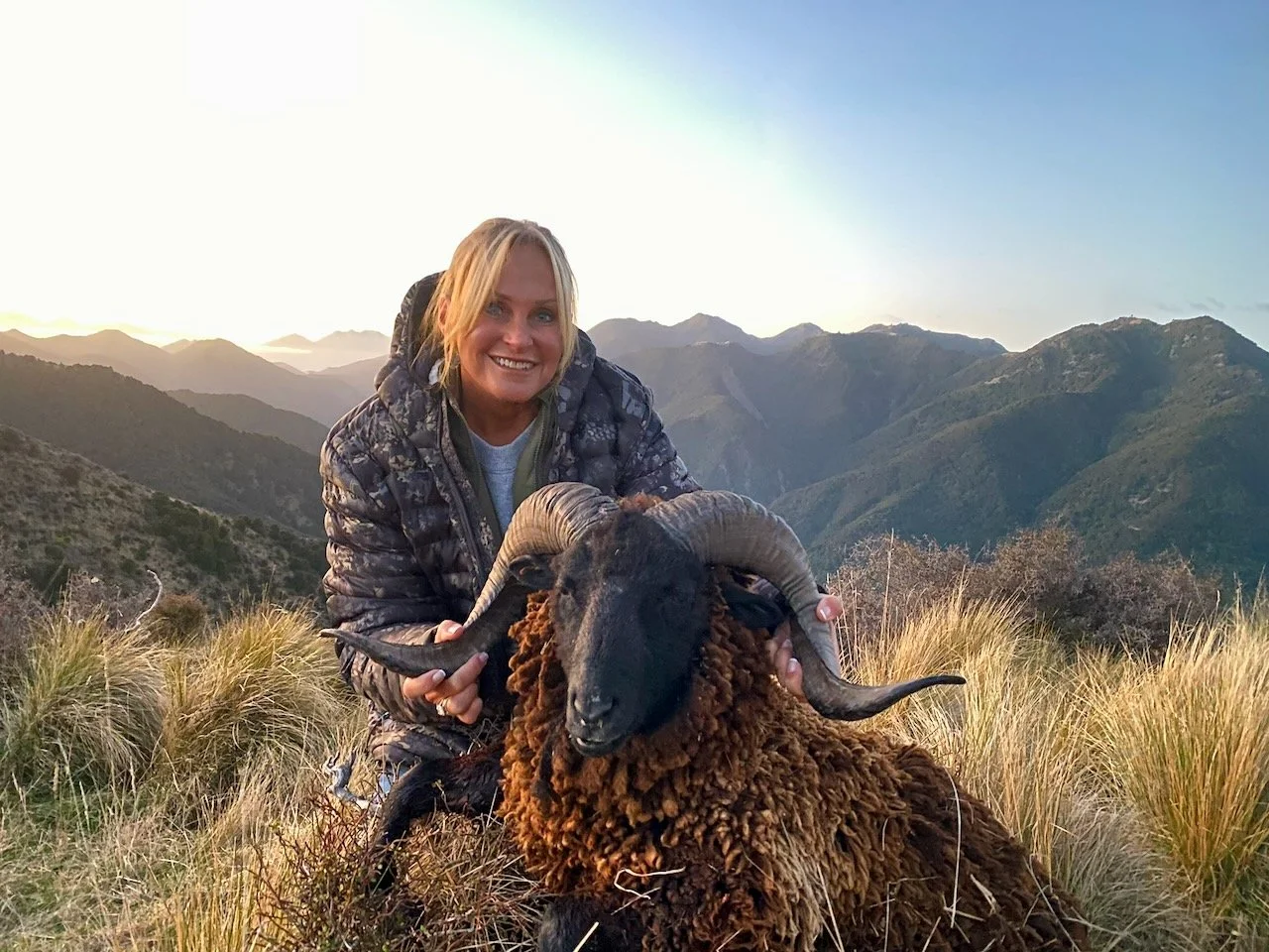 A woman holding a large black sheep with curved horns on a mountain trail during sunset.