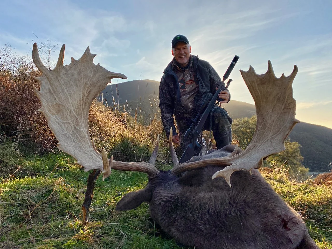 A man smiling outdoors, kneeling next to a large dead elk with big antlers, holding a rifle, with mountains and a blue sky in the background.