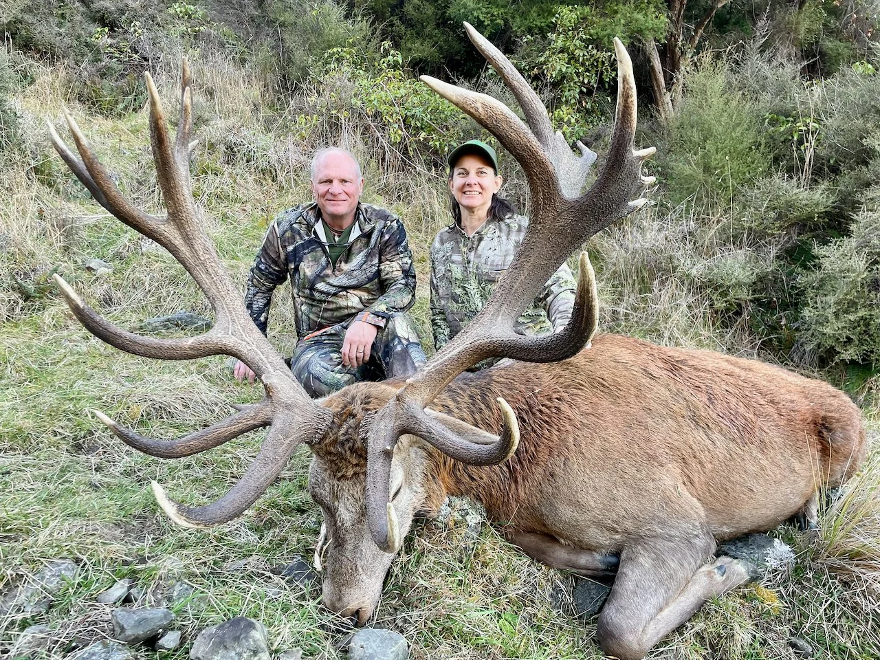 Two hunters kneeling behind a large dead elk with impressive antlers, surrounded by grass and shrubs.