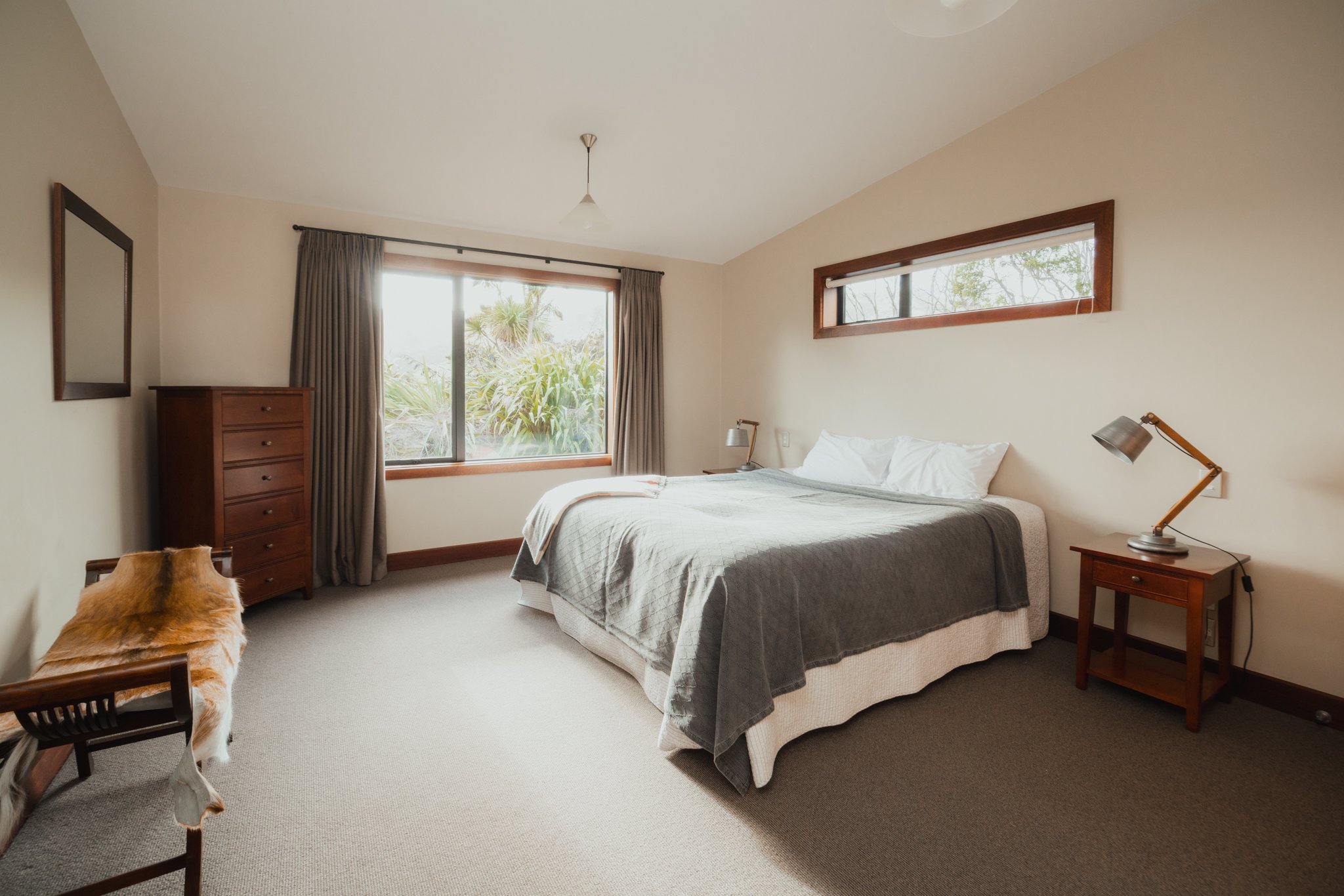 A bedroom with beige walls, a double bed with gray and white bedding, two wooden nightstands with lamps, a tall chest of drawers, a rectangular mirror, a window with beige curtains, and a smaller horizontal window above the bed.