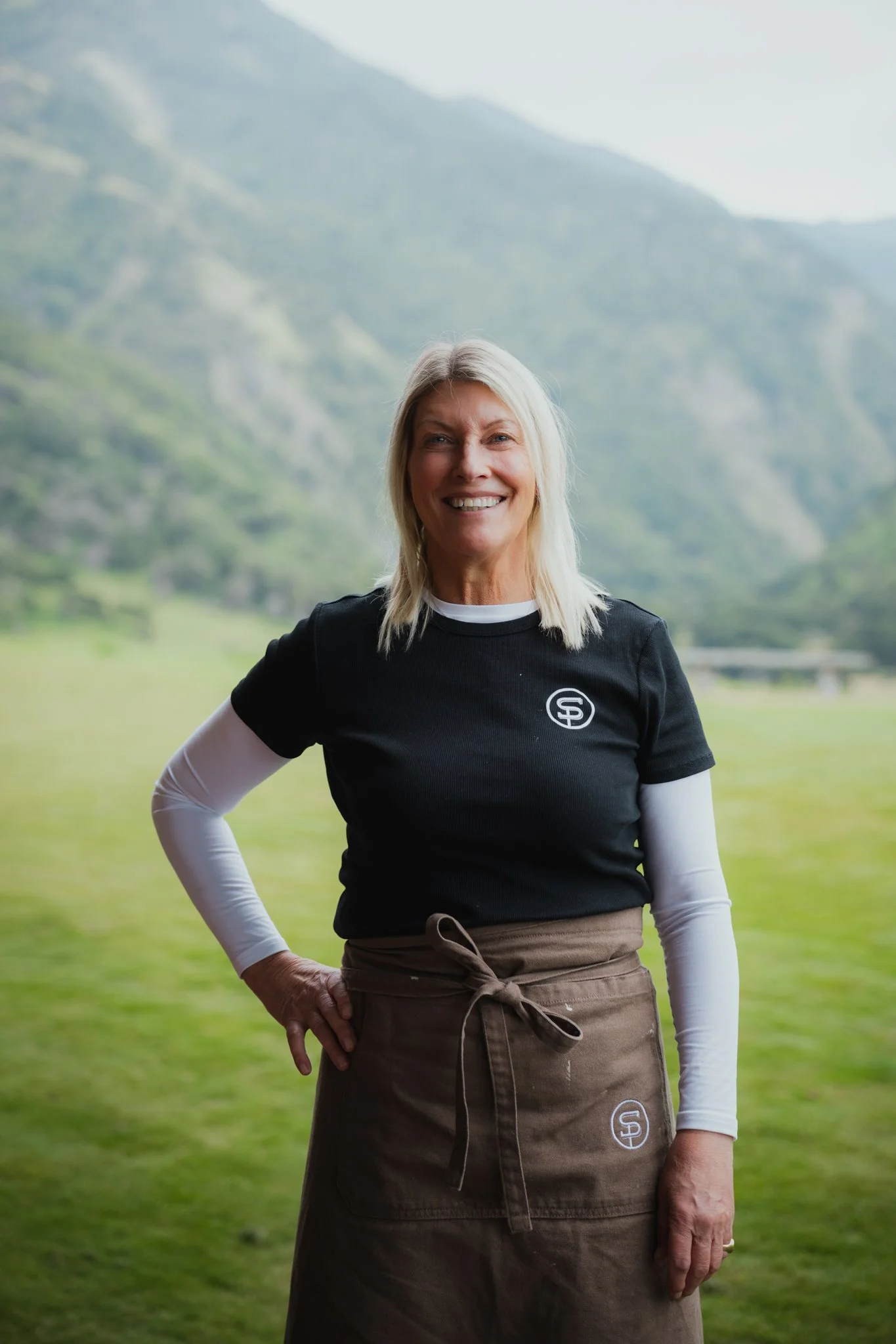 A woman smiling outdoors. She is wearing a black T-shirt with a logo, a white long-sleeve shirt underneath, and a brown apron tied at the waist. The background features mountains and greenery.