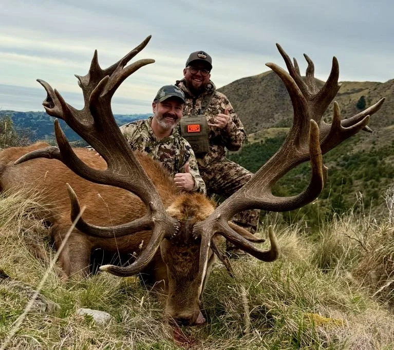 Two men in camouflage hunting gear posing with a large elk with big antlers. The elk is lying on the grass in a hilly, open landscape.