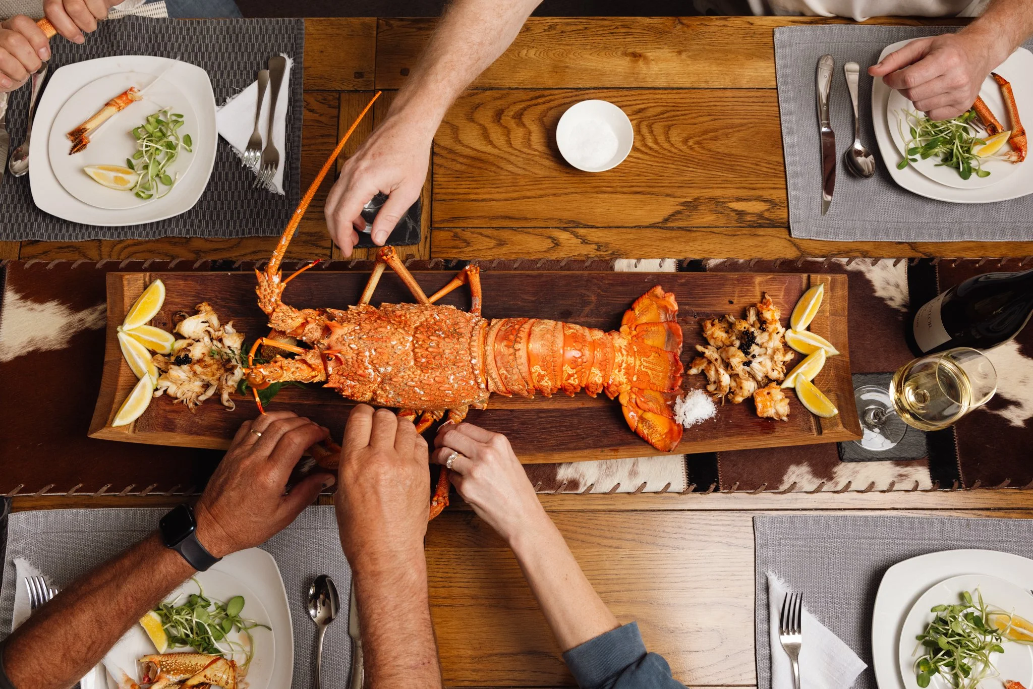 Multiple hands reaching for a large cooked lobster on a wooden platter. There are lemon wedges and some seafood scraps around the lobster. The table has plates with salad and silverware.