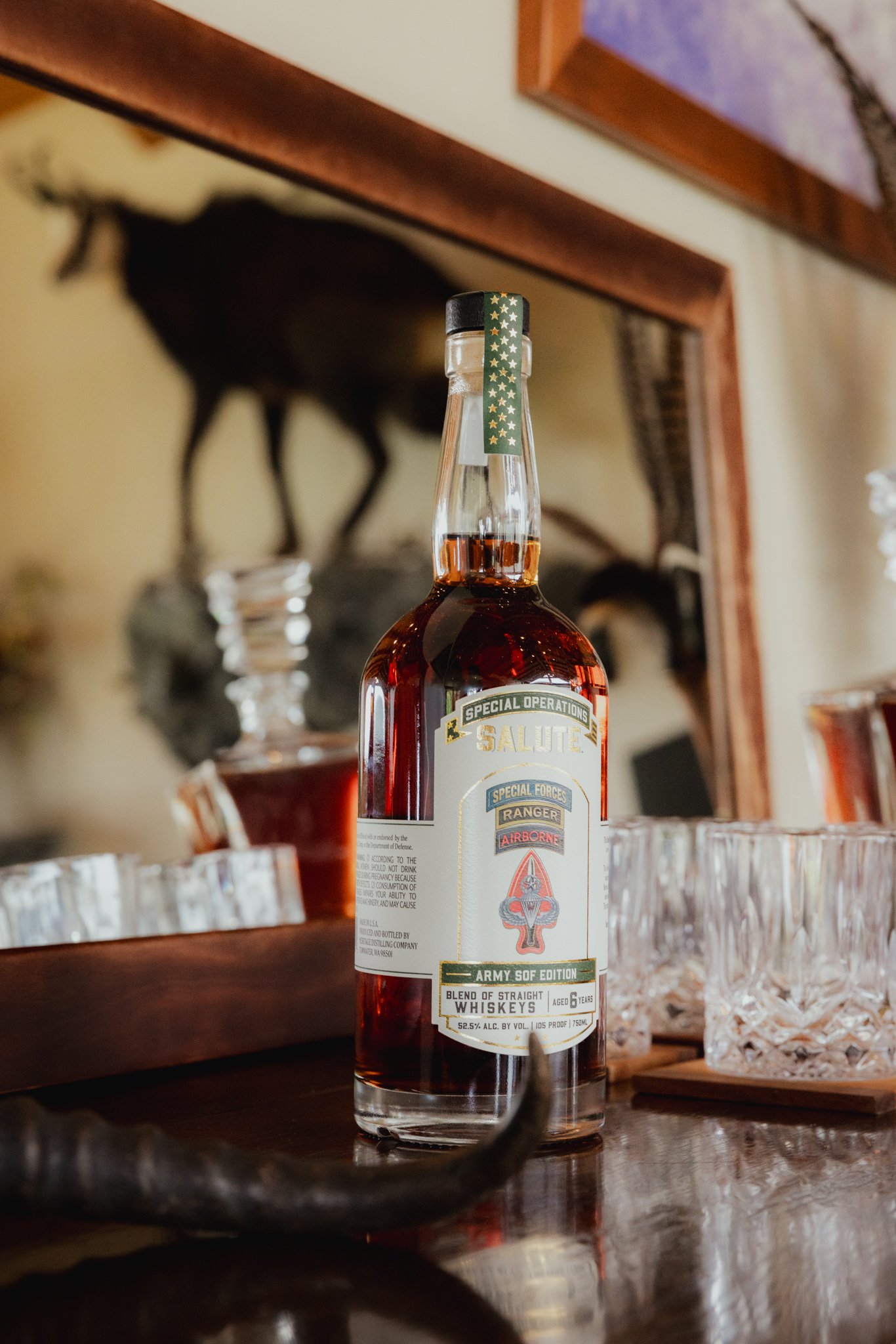Close-up of a whiskey bottle with a blurred background of a mounted animal and glassware on a wooden surface.