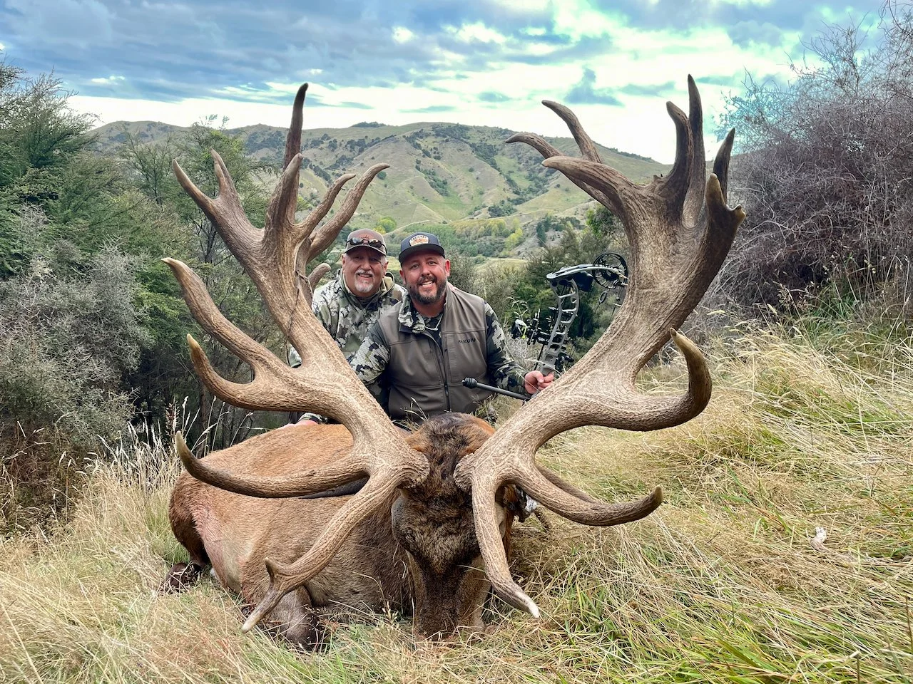 Two men posing behind a large elk with impressive antlers in a grassy field with hills and trees in the background.