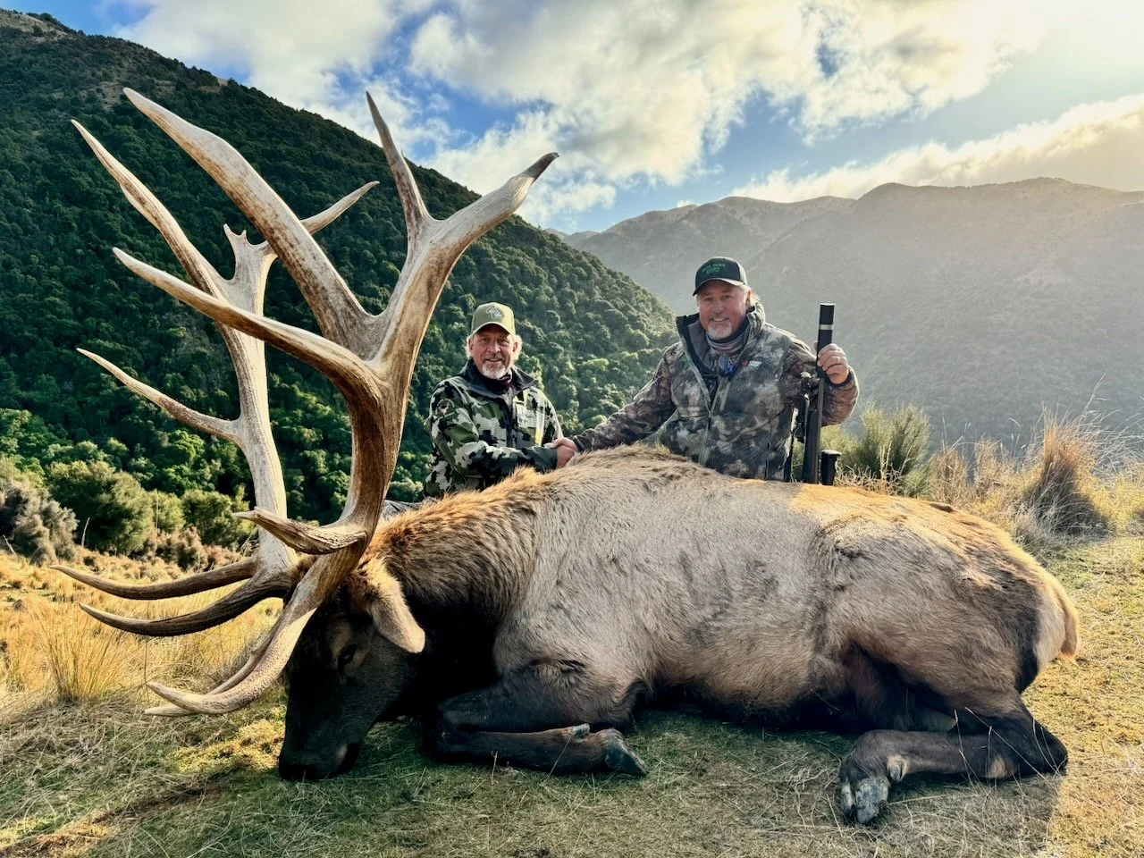 Two men in camouflage clothing after a successful elk hunt, shaking hands behind a large dead elk with impressive antlers, in a mountainous landscape with green trees and a partly cloudy sky.