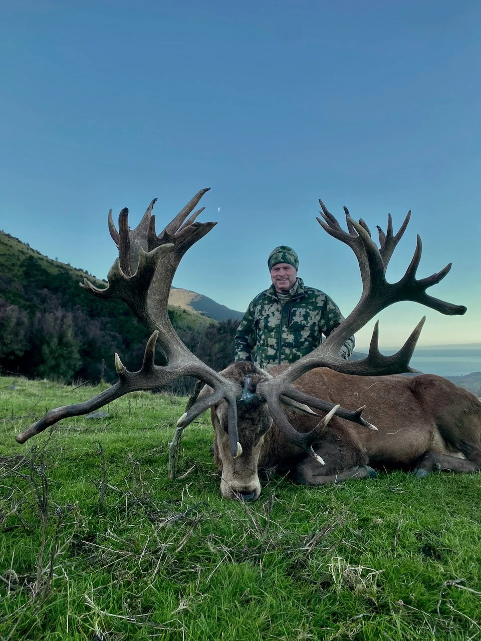 A man in camouflage clothing stands behind a large elk with impressive antlers lying on the grass, with hills and a partly cloudy sky in the background.