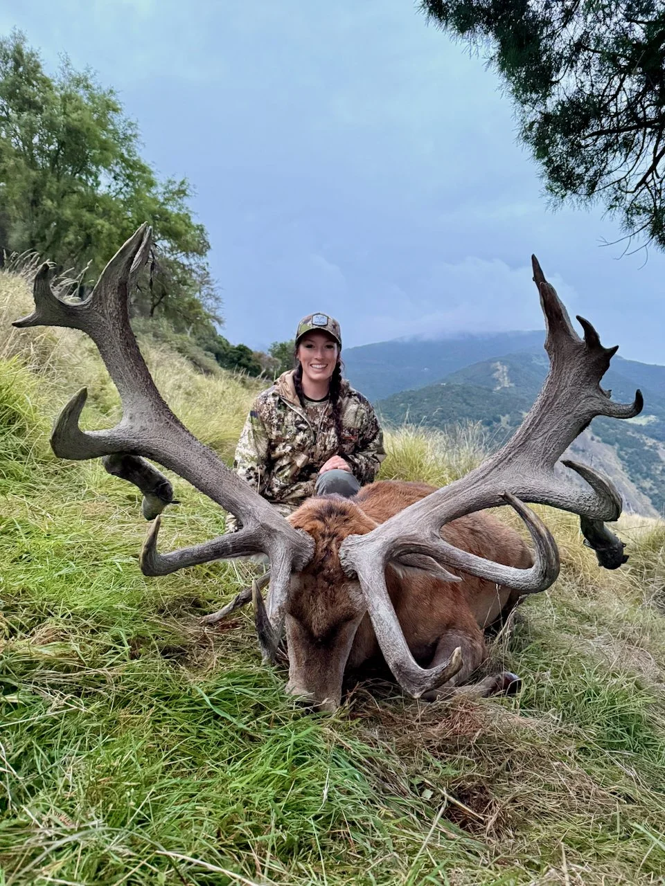 A woman dressed in camouflage hunting gear kneels behind a large elk with impressive antlers in a grassy field with trees and mountains in the background.