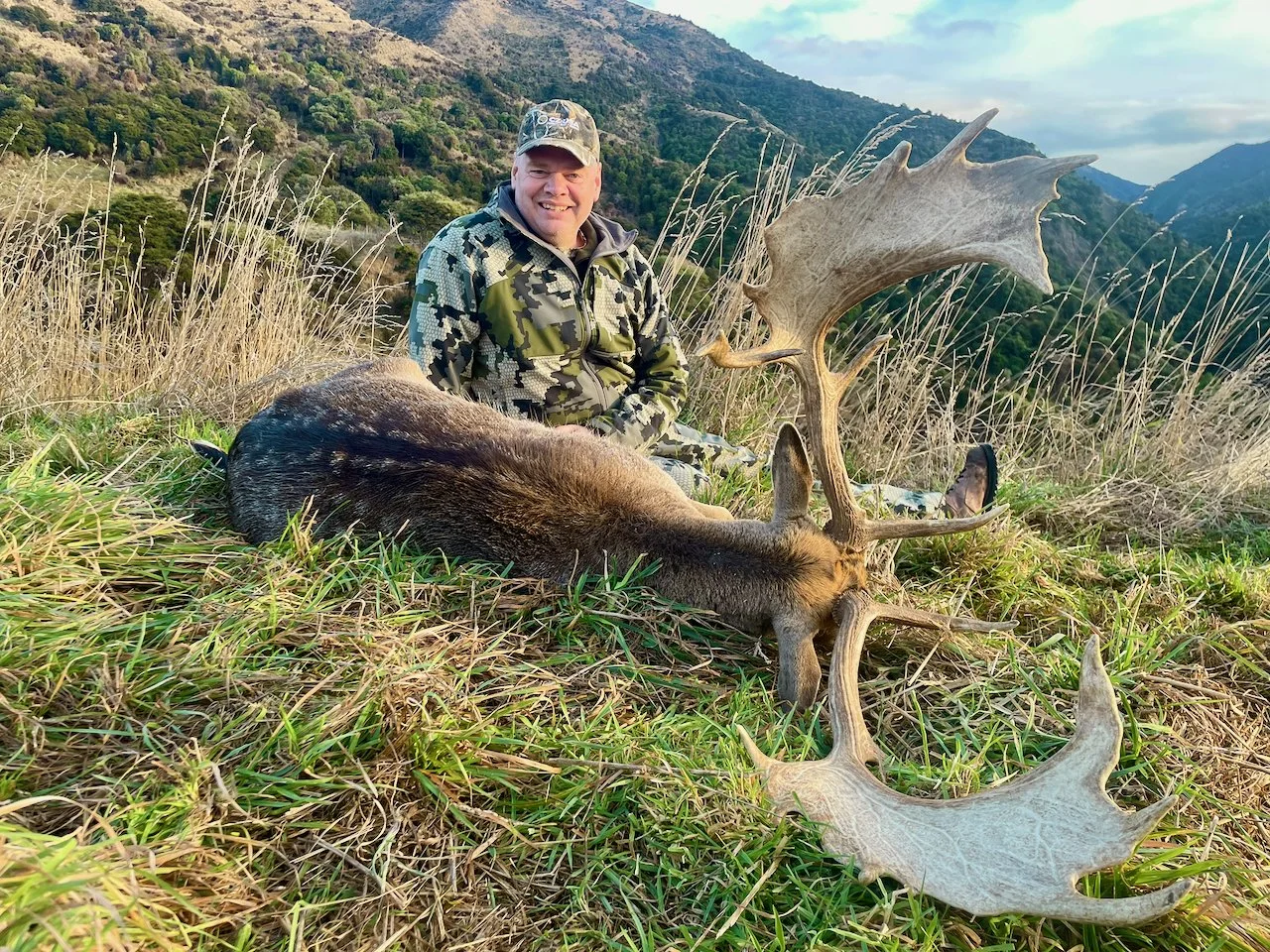 A man dressed in camouflage clothing sitting on grass in a mountainous landscape with tall grasses, smiling at the camera, next to a large slain elk with massive antlers.