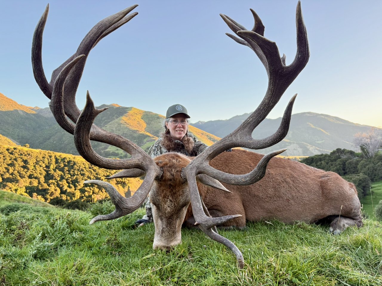 Woman in camouflage clothing and cap sitting behind a large elk with big antlers in a grassy field with mountains in the background.