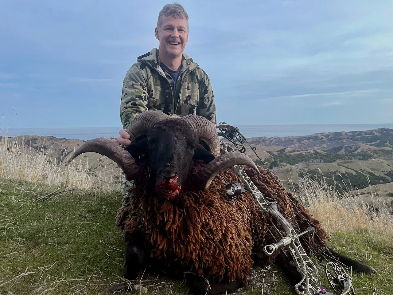 Man smiling and holding a harvested ram with large curved horns, sitting on grass in a hilly landscape with overcast sky.
