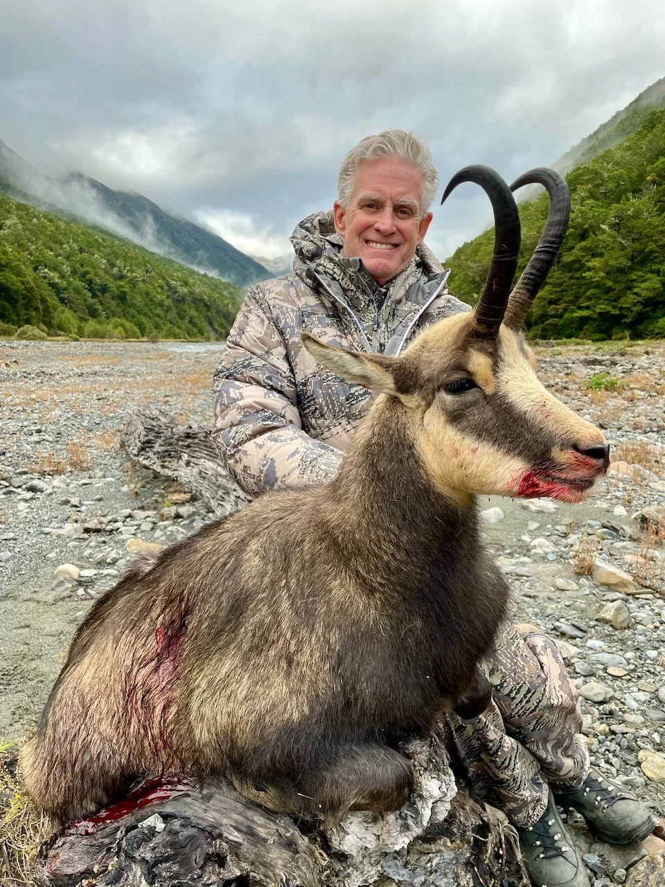 A man in camouflage outdoor clothing sitting outdoors with a slain goat-like animal with long, curved horns, on a rocky terrain with mountains and cloudy sky in the background.