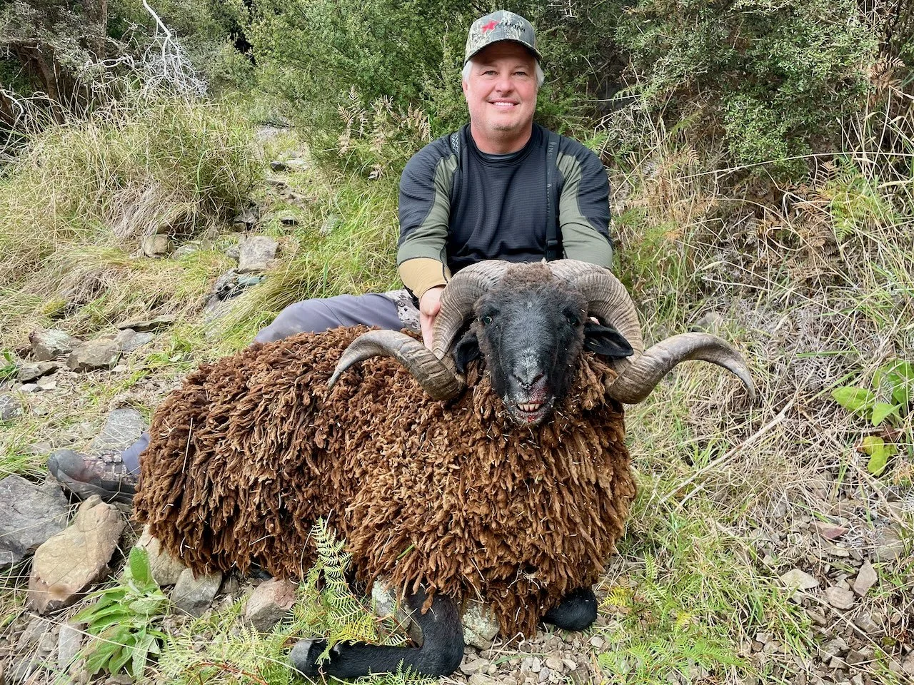 A man in outdoor clothing and a camouflage cap smiling while holding a large ram with thick, curly horns and a shaggy brown coat in a grassy, rocky natural setting.