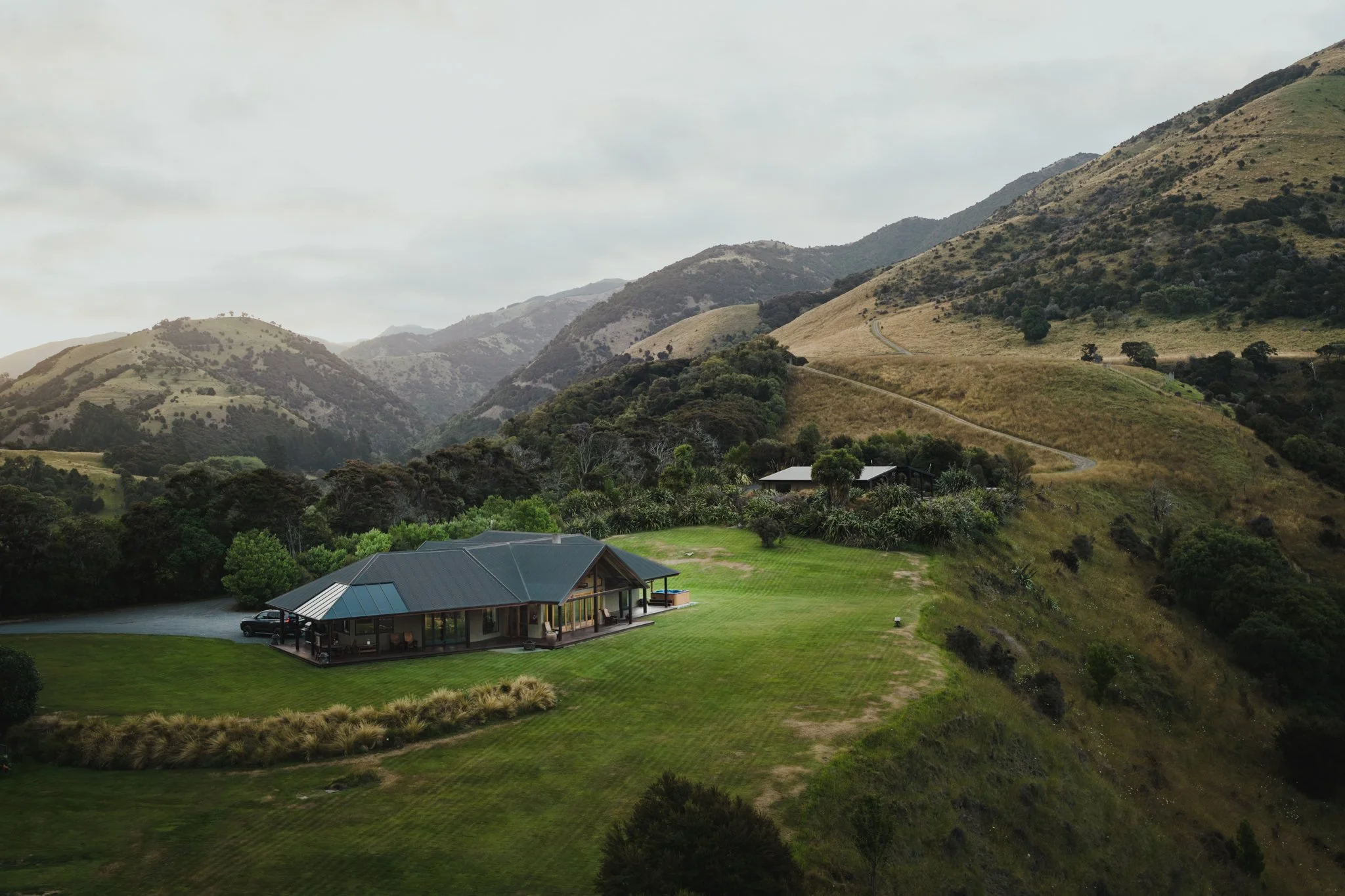 A house with a blue metal roof and wraparound porch situated on a large grassy lawn, surrounded by trees, with rolling green hills and mountains in the background under a cloudy sky.
