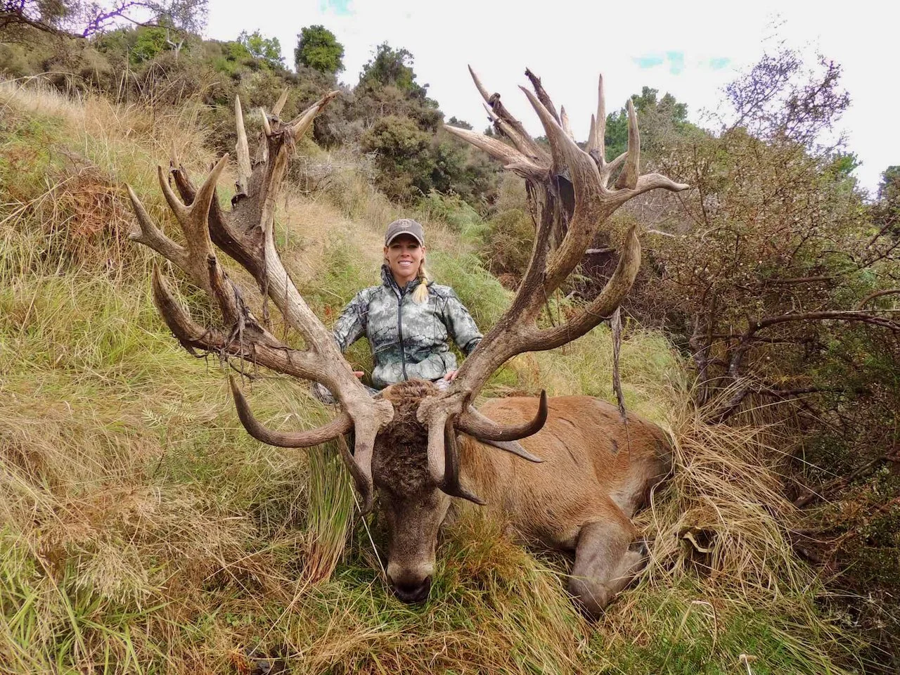 A woman in camouflage jacket and cap sitting behind a large elk with impressive antlers on a grassy hillside with trees and shrubs in the background.