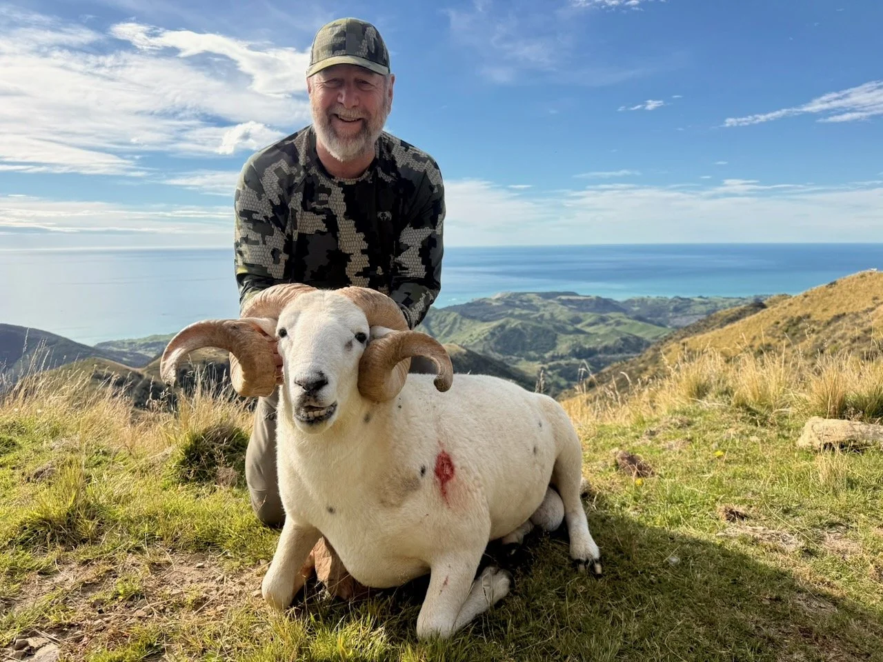 A smiling man in camouflage clothing kneels behind a white sheep with large curled horns, sitting on a grassy hillside with a scenic view of mountains and the ocean in the background.
