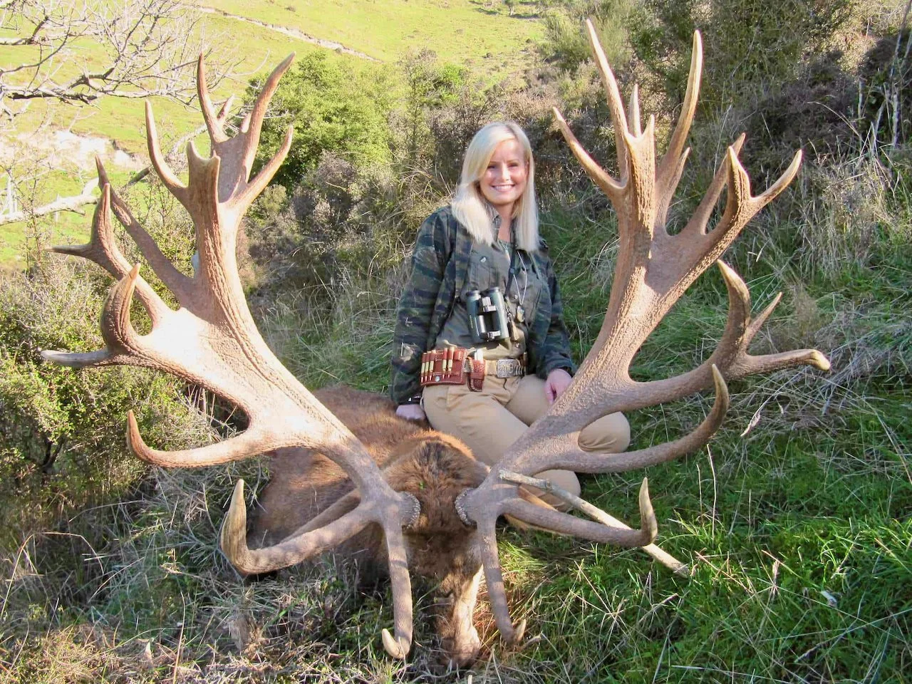 A woman with blonde hair kneeling next to a large elk with impressive antlers in a grassy outdoor setting.
