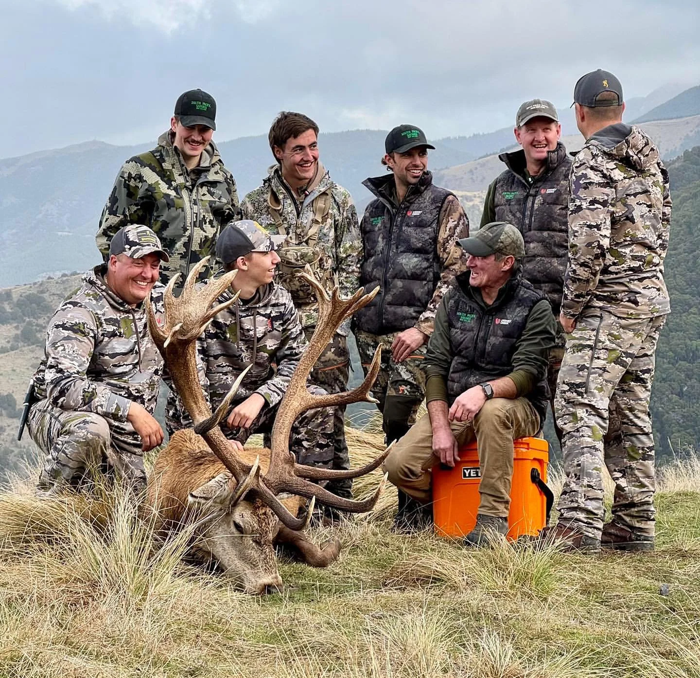 Group of people in camouflage clothing posing with a large, freshly hunted deer with antlers, outdoors with mountainous background.