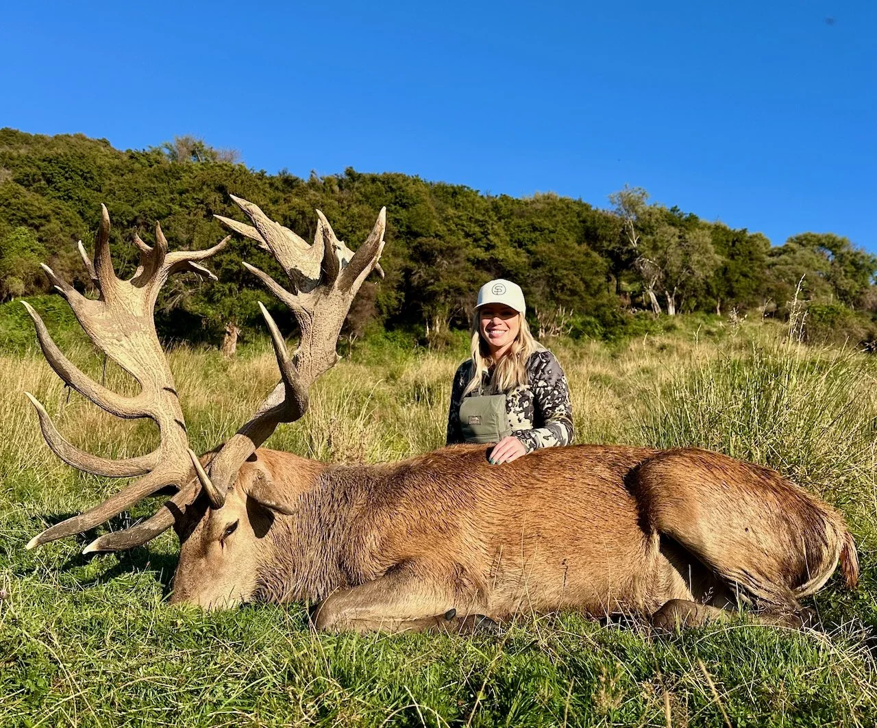 A woman is kneeling next to a large elk with impressive antlers, lying on the grass in a field with trees and a blue sky in the background.