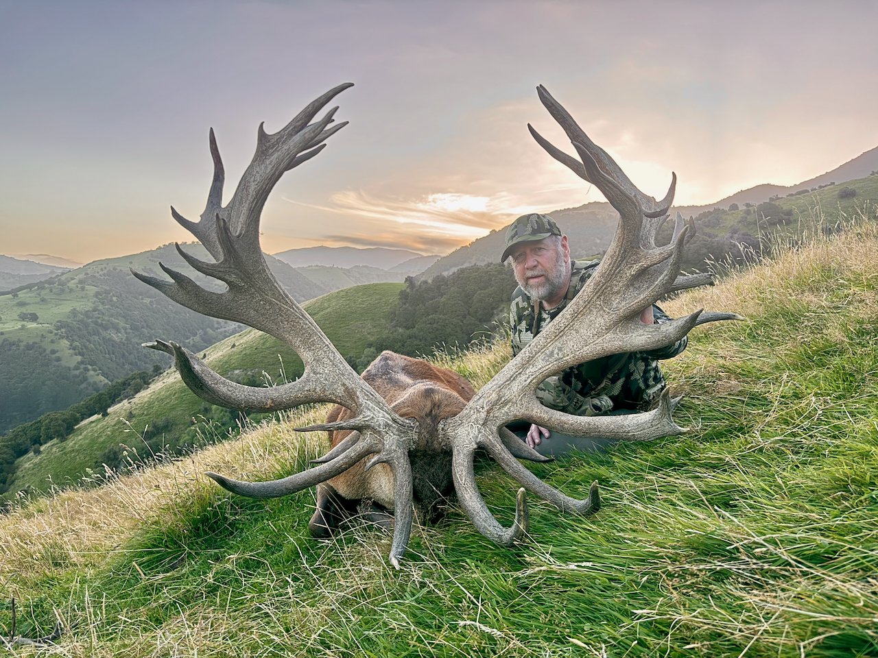 A man in camouflage clothing kneels next to a large deer with massive antlers on a grassy hillside at sunset with rolling hills in the background.