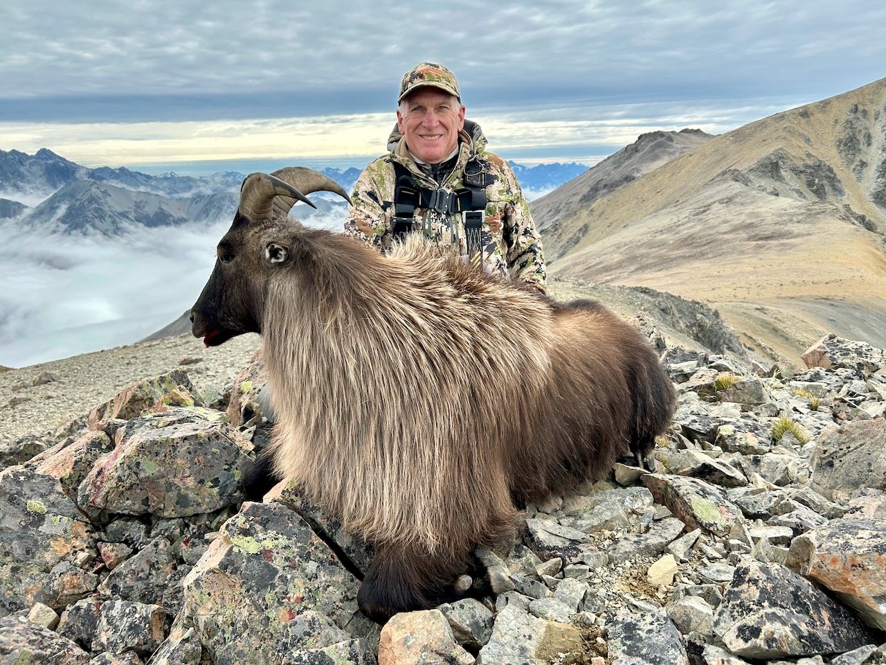 A man in outdoor camouflage gear and a cap on rocky mountain terrain, smiling, with a large goat with curved horns in front of him. Mountains and cloudy sky in the background.