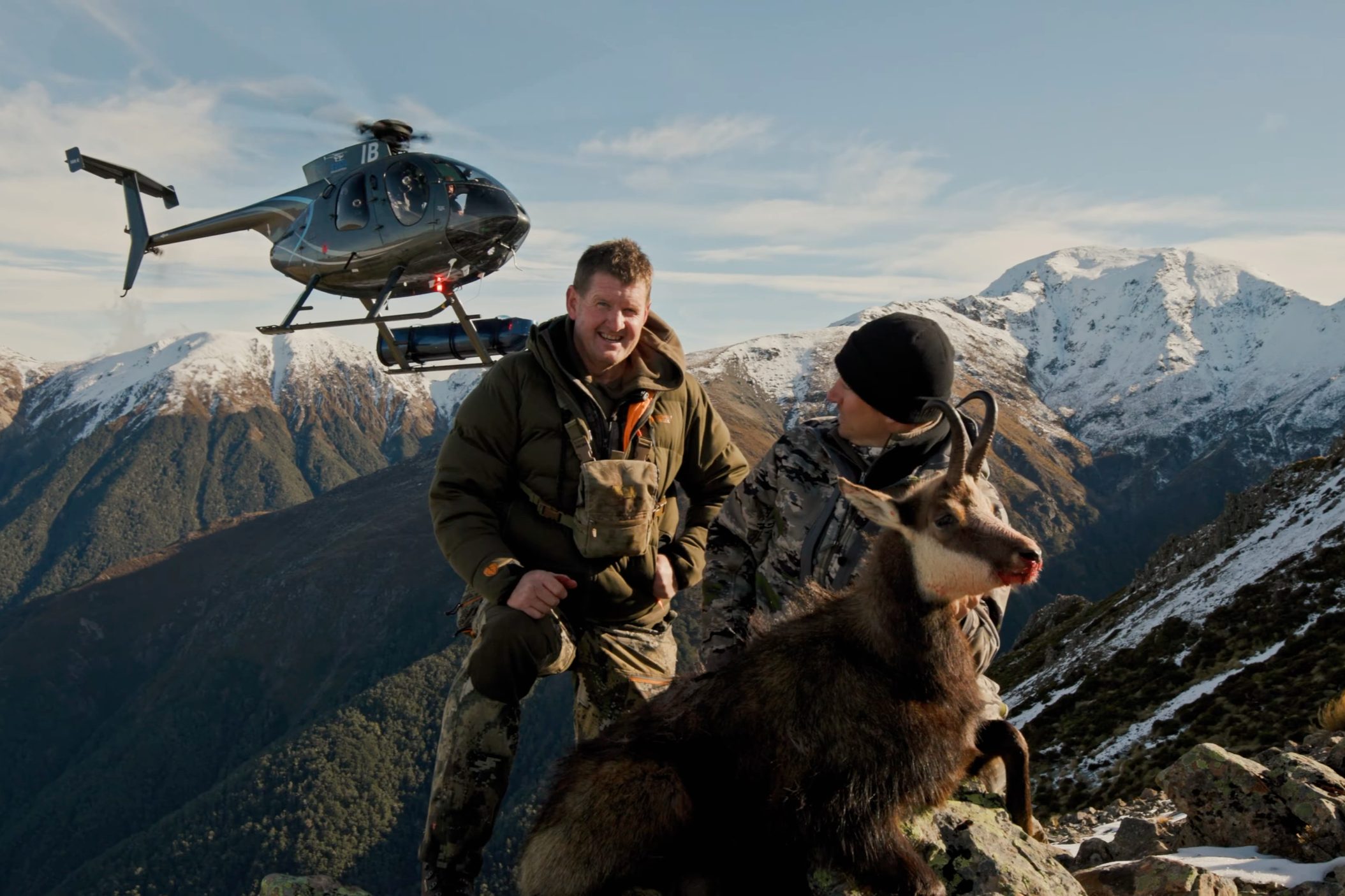 Two men with backpacks, a chamois buck, and a helicopter on a mountain with snow-capped peaks in the background.