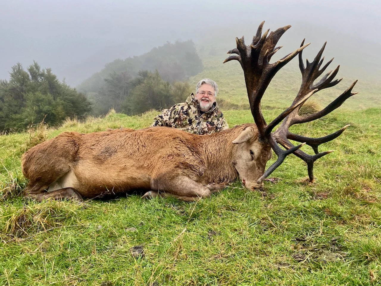 A man smiling and lying behind a large elk with big antlers, on a grassy hillside with foggy trees in the background.