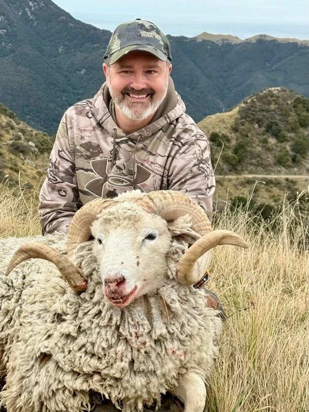 A man outdoors in a grassy field, smiling and posing with a sheep that has large, curved horns, set against a backdrop of rolling hills and mountains.