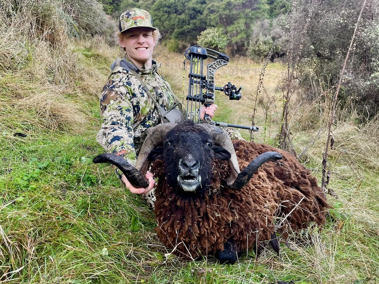 A young man in camouflage hunting gear is smiling and holding a large, black sheep-like animal with curved horns in a grassy, wooded area.
