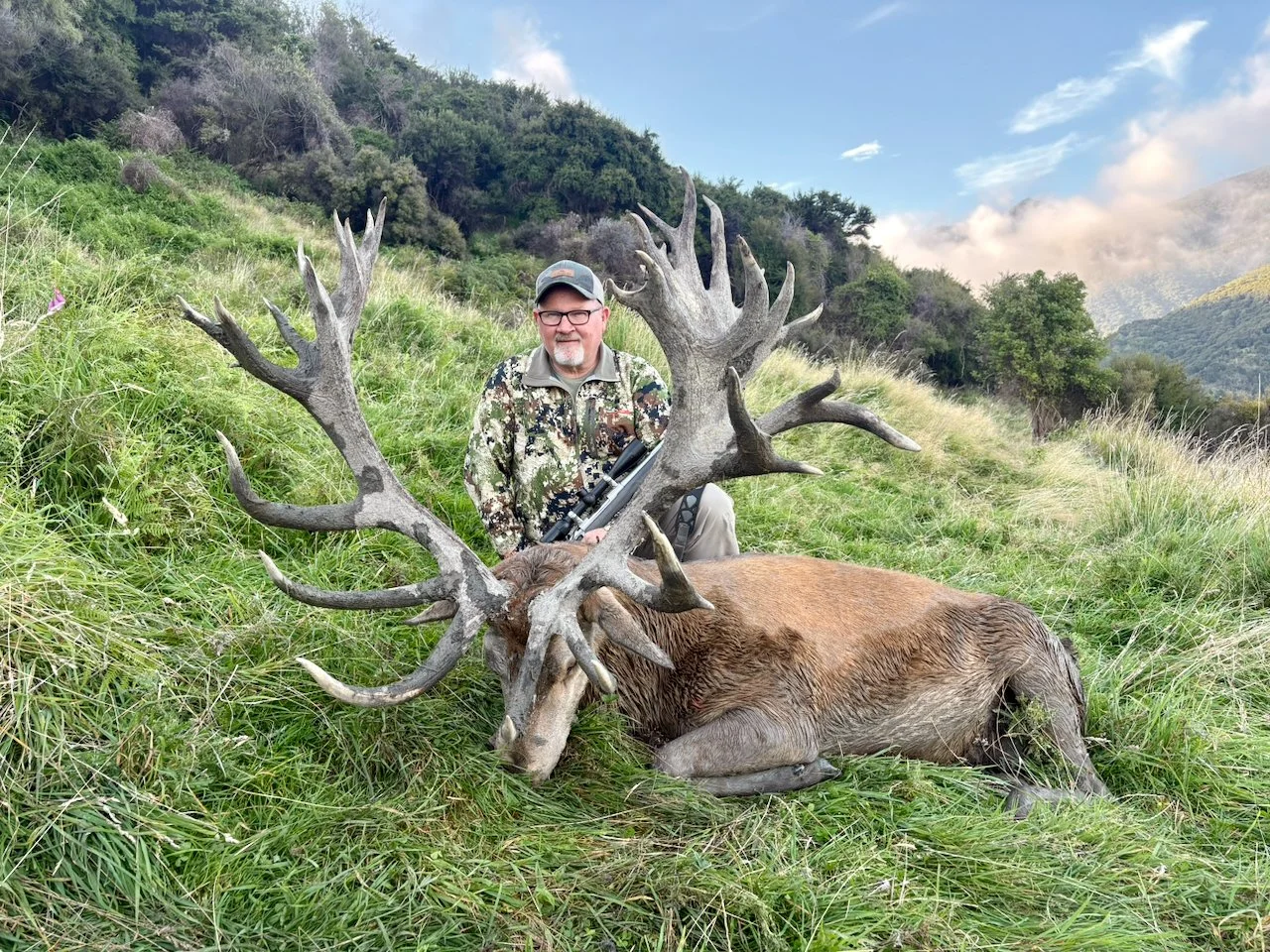 A man kneeling on grassy terrain next to a large elk with impressive antlers, holding a rifle, in an outdoor mountain landscape with rolling hills, trees, and a partly cloudy sky.