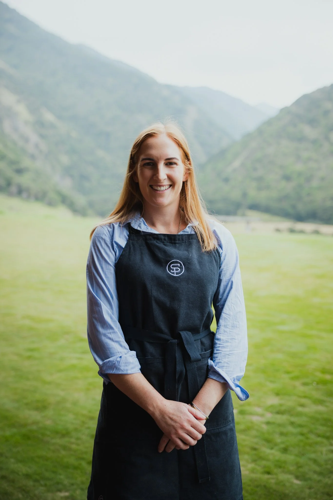 A woman with blonde hair wearing a blue shirt and black apron standing outdoors in a green valley surrounded by mountains.
