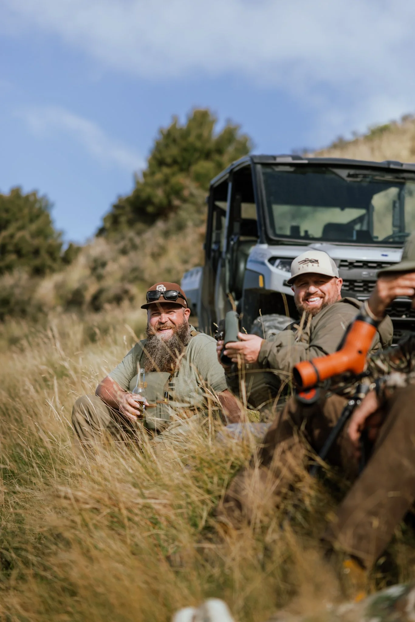Two men sitting in tall grass outdoors with a utility vehicle in the background, smiling and taking a selfie, one holding a drink, under a blue sky with some clouds.