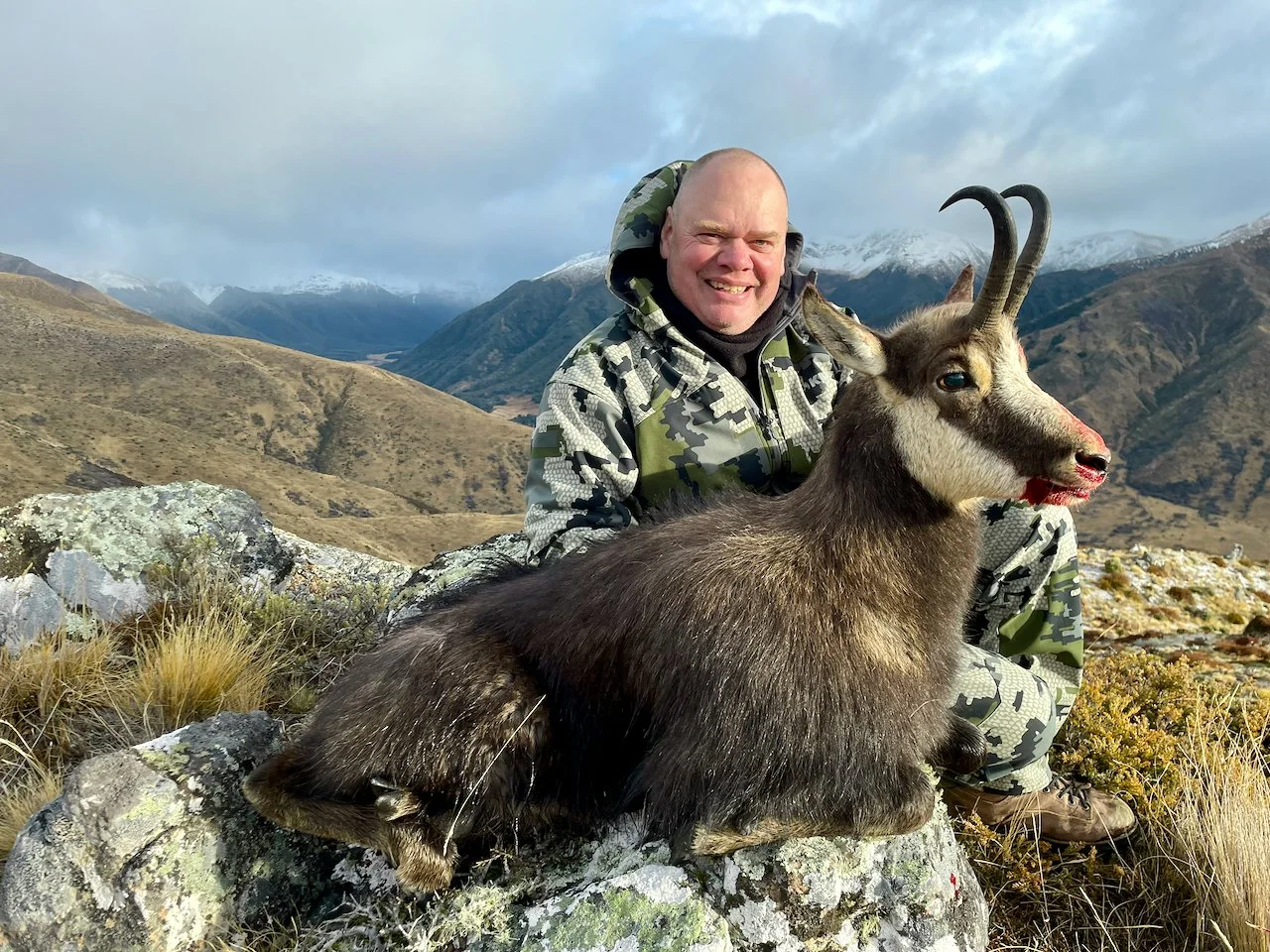 Man in camouflage jacket posing with a mountain goat in a mountainous landscape with snow-capped peaks in the background.