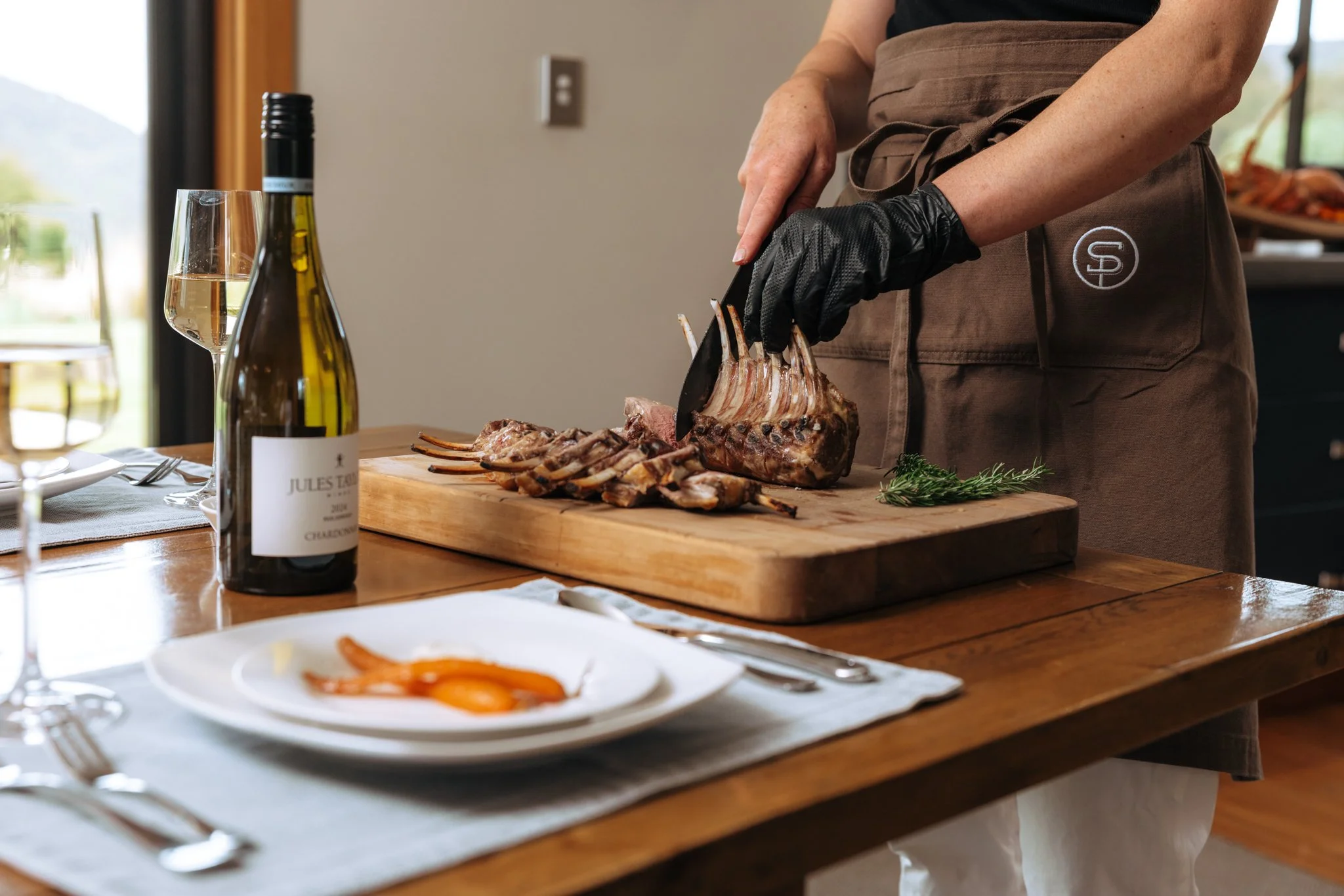 Person slicing cooked rack of lamb on wooden cutting board with glass of white wine and bottle on dining table, with plates of food in foreground.
