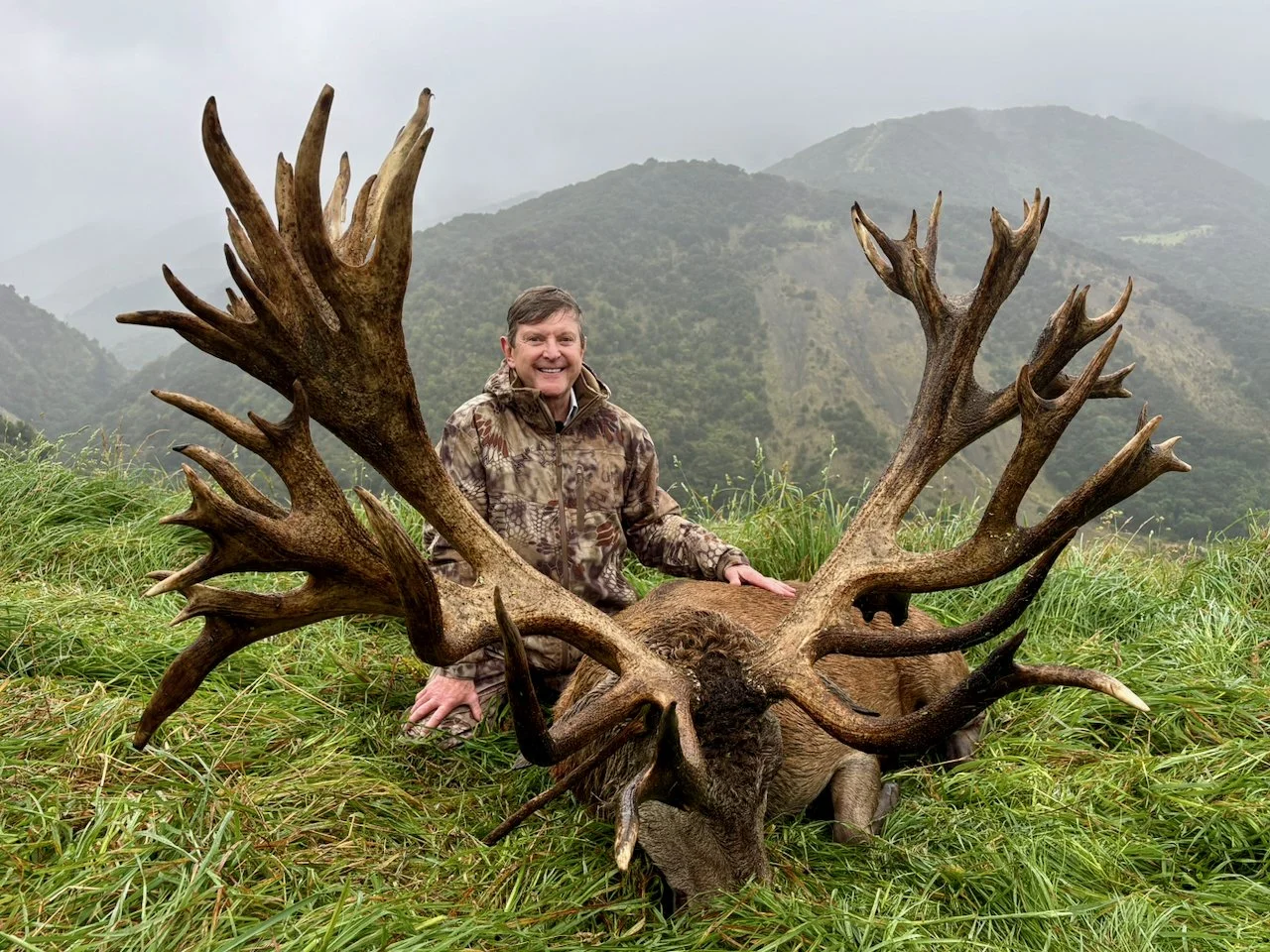 A man in camouflage jacket kneeling on grass with a large, dead elk with big antlers in a mountainous outdoor landscape.