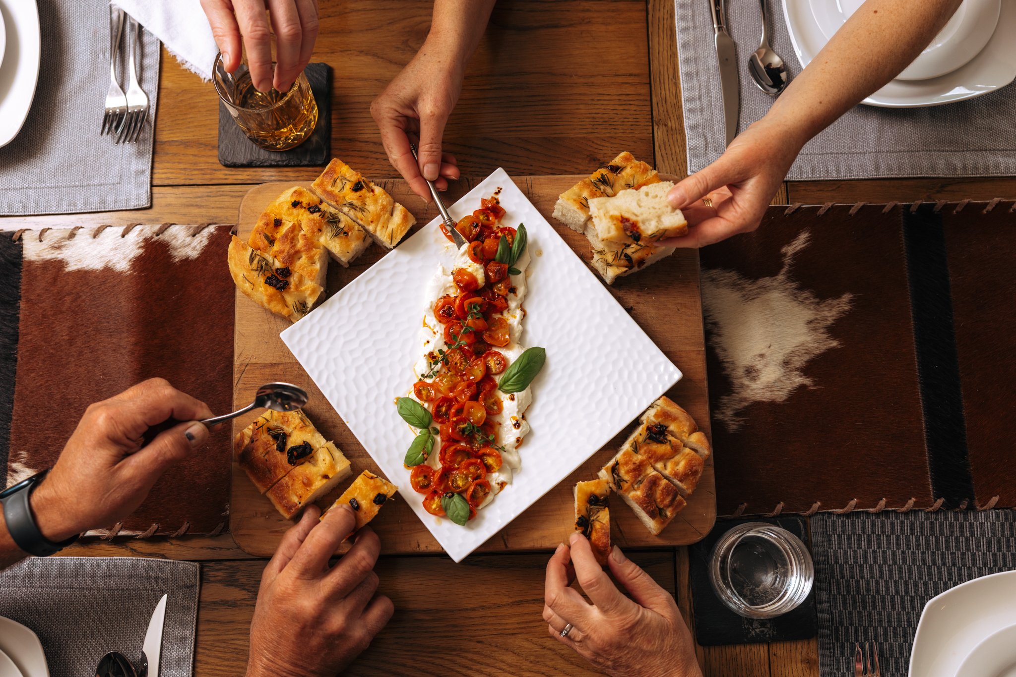 People sharing slices of focaccia bread with cherry tomato and basil topping at a dining table.