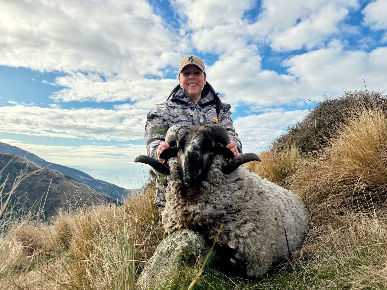 A woman wearing a camouflage jacket and a light-colored cap is kneeling on a grassy hillside, holding a large ram with thick curly wool and prominent curved horns. The background features mountains and a partly cloudy sky.