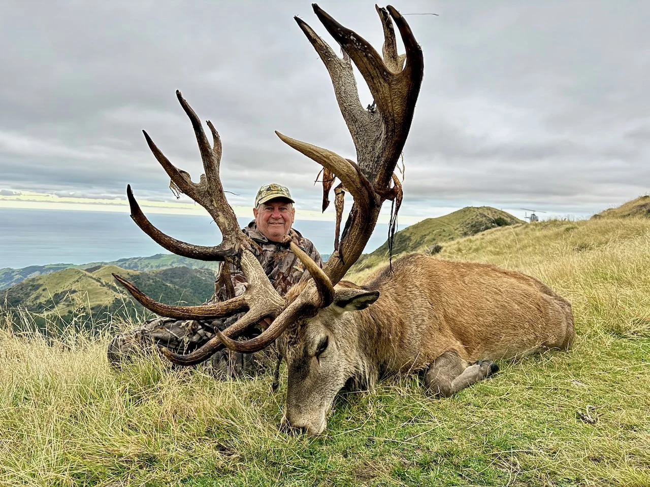 A hunter in camouflage clothing posing with a large fallen deer with massive antlers on a grassy hillside under a cloudy sky.